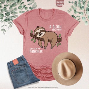 K&ouml;nnte beinhalten: Mauvefarbenes T-Shirt mit einem Faultier-Motiv auf einem Ast und dem Text "A sloth does more work than my pancreas". Die Abbildung zeigt auch eine blaue Jeans und einen beigen Hut.