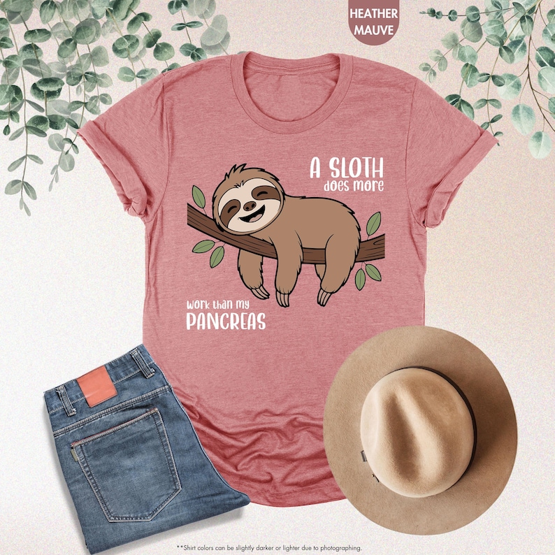 K&ouml;nnte beinhalten: Mauvefarbenes T-Shirt mit einem Faultier-Motiv auf einem Ast und dem Text "A sloth does more work than my pancreas". Die Abbildung zeigt auch eine blaue Jeans und einen beigen Hut.
