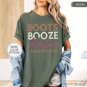 Pu&ograve; includere: T-shirt verde muschio con la scritta "BOOTS BOOZE & Besties" in marrone, bianco e rosa. Il testo "NASHVILLE, TN" &egrave; stampato sotto. La maglietta ha le maniche arrotolate ed &egrave; abbinata a una giacca di jeans.