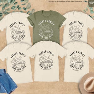 May include: Several t-shirts in cream and olive green, featuring a black graphic. The design reads "PARKER FAMILY ROAD TRIP 2026" with a mountain and lake illustration. The image also includes blue jeans and a hat.