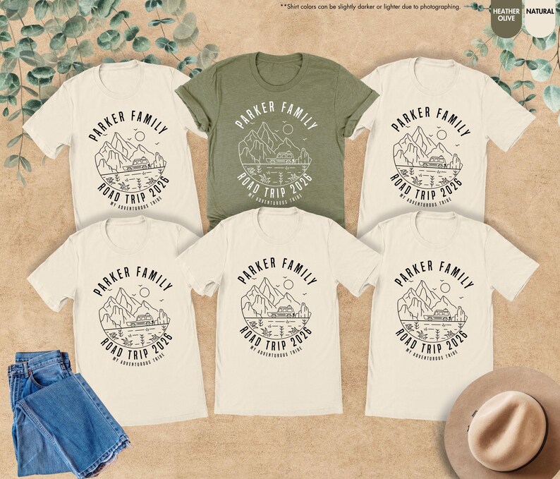 May include: Several t-shirts in cream and olive green, featuring a black graphic. The design reads "PARKER FAMILY ROAD TRIP 2026" with a mountain and lake illustration. The image also includes blue jeans and a hat.