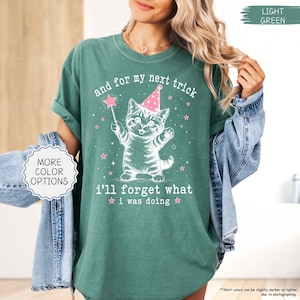 May include: A light green short-sleeve t-shirt featuring a white graphic of a kitten wearing a pink party hat and holding a star wand. The text reads, and for my next trick i'll forget what i was doing, surrounded by small pink stars.