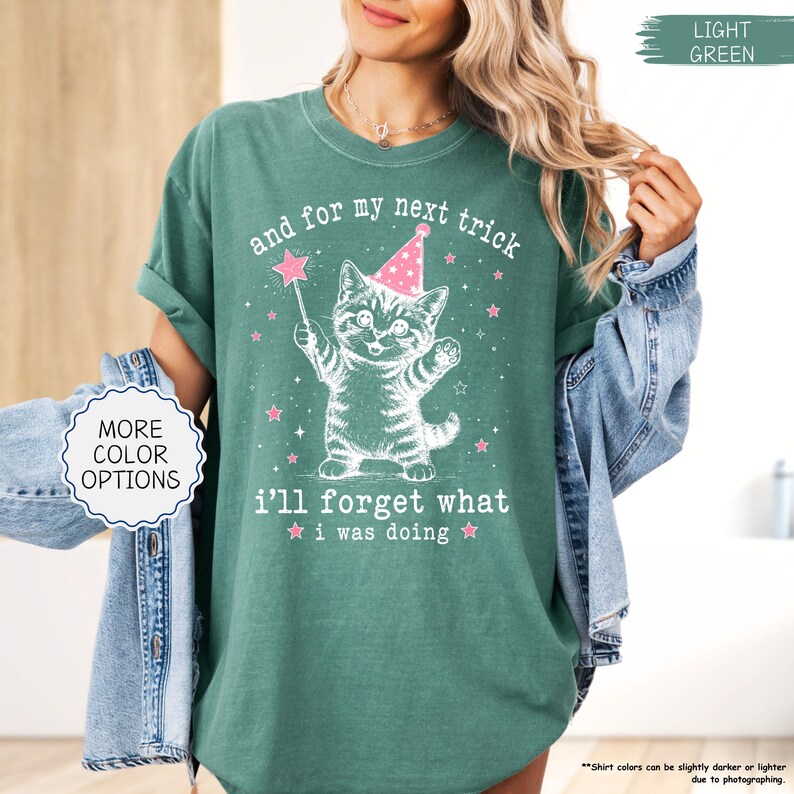 May include: A light green short-sleeve t-shirt featuring a white graphic of a kitten wearing a pink party hat and holding a star wand. The text reads, and for my next trick i'll forget what i was doing, surrounded by small pink stars.