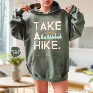 K&ouml;nnte beinhalten: Milit&auml;rgr&uuml;ner Kapuzenpullover mit dem wei&szlig;en Text "TAKE A HIKE." Das Design umfasst eine Bergkette und B&auml;ume. Der Hoodie hat eine Vordertasche und eine Kapuze mit Kordelzug.