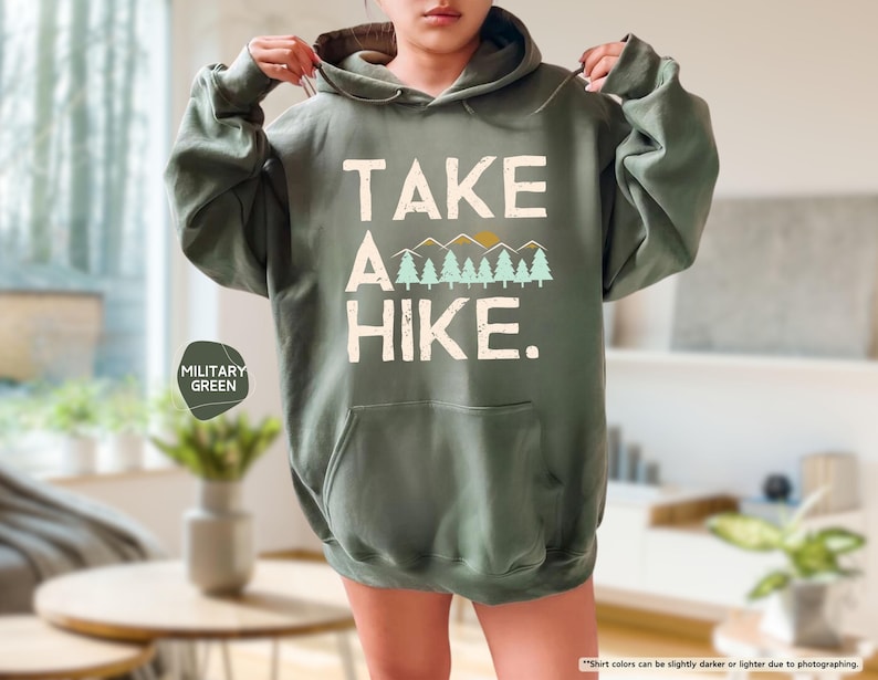K&ouml;nnte beinhalten: Milit&auml;rgr&uuml;ner Kapuzenpullover mit dem wei&szlig;en Text "TAKE A HIKE." Das Design umfasst eine Bergkette und B&auml;ume. Der Hoodie hat eine Vordertasche und eine Kapuze mit Kordelzug.