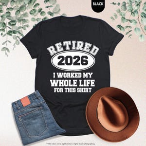 Puede incluir: Camiseta negra con el texto "RETIRED 2026" encima de "I WORKED MY WHOLE LIFE FOR THIS SHIRT" en blanco. La camiseta se combina con unos vaqueros azules y un sombrero marr&oacute;n. La palabra "BLACK" est&aacute; en la esquina superior derecha.
