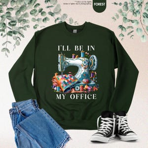 Pode incluir: Uma camisa de frio verde escura com o texto "I'll be in my office" e uma ilustra&ccedil;&atilde;o colorida de uma m&aacute;quina de costura com tecido e linha.