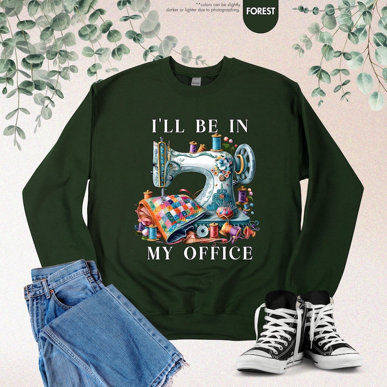 Pode incluir: Uma camisa de frio verde escura com o texto "I'll be in my office" e uma ilustra&ccedil;&atilde;o colorida de uma m&aacute;quina de costura com tecido e linha.
