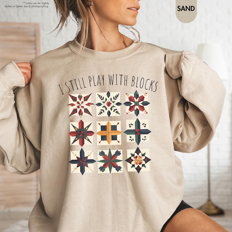 May include: A beige sweatshirt with the text "I still play with blocks" and a pattern of colorful quilt blocks.