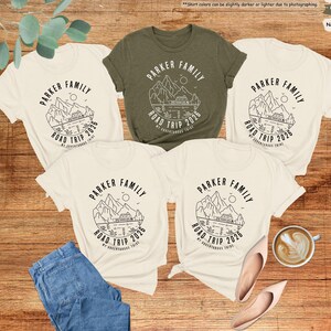 May include: Five t-shirts with a family road trip design. The shirts are in natural and olive green colours. The design includes the text "PARKER FAMILY ROAD TRIP 2026" with a mountain and lake graphic. A pair of jeans, shoes, and a coffee cup are also in the image.
