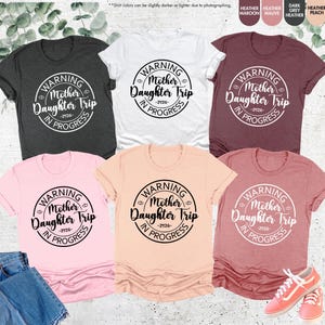 K&ouml;nnte beinhalten: Eine Auswahl an T-Shirts in verschiedenen Farben, darunter Grau, Wei&szlig; und Rosa. Jedes Shirt zeigt eine runde Grafik mit dem Text "WARNING Mother Daughter Trip IN PROGRESS -2026-".