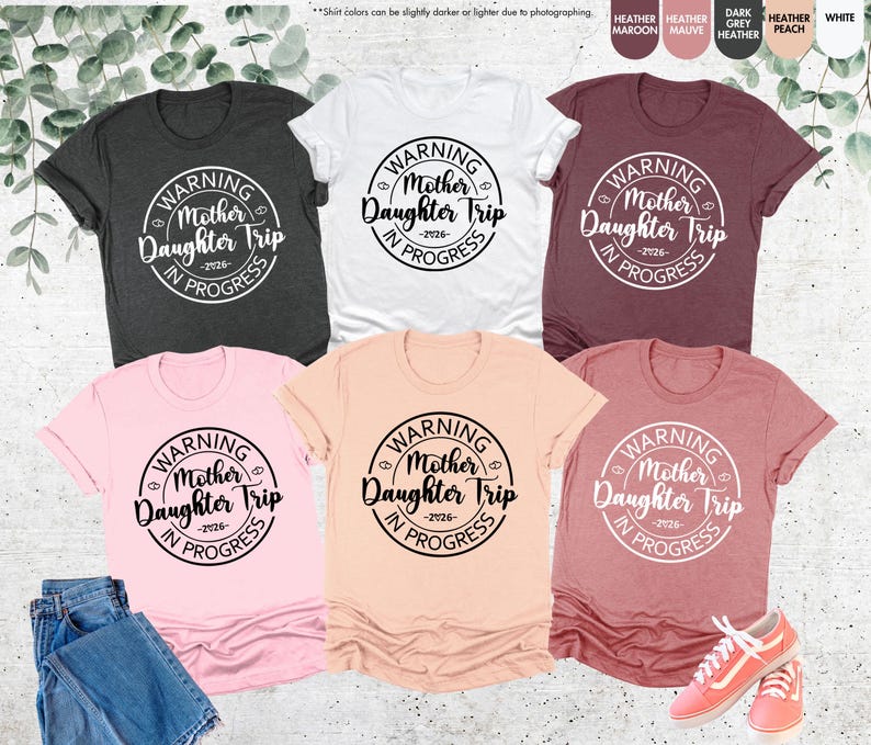 K&ouml;nnte beinhalten: Eine Auswahl an T-Shirts in verschiedenen Farben, darunter Grau, Wei&szlig; und Rosa. Jedes Shirt zeigt eine runde Grafik mit dem Text "WARNING Mother Daughter Trip IN PROGRESS -2026-".