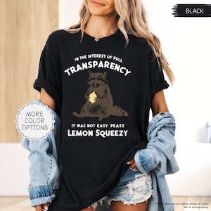 May include: Black t-shirt with a raccoon graphic holding a lemon. The text on the shirt reads "IN THE INTEREST OF FULL TRANSPARENCY", "IT WAS NOT EASY PEASY", and "LEMON SQUEEZY". The shirt also has a "MORE COLOR OPTIONS" button.