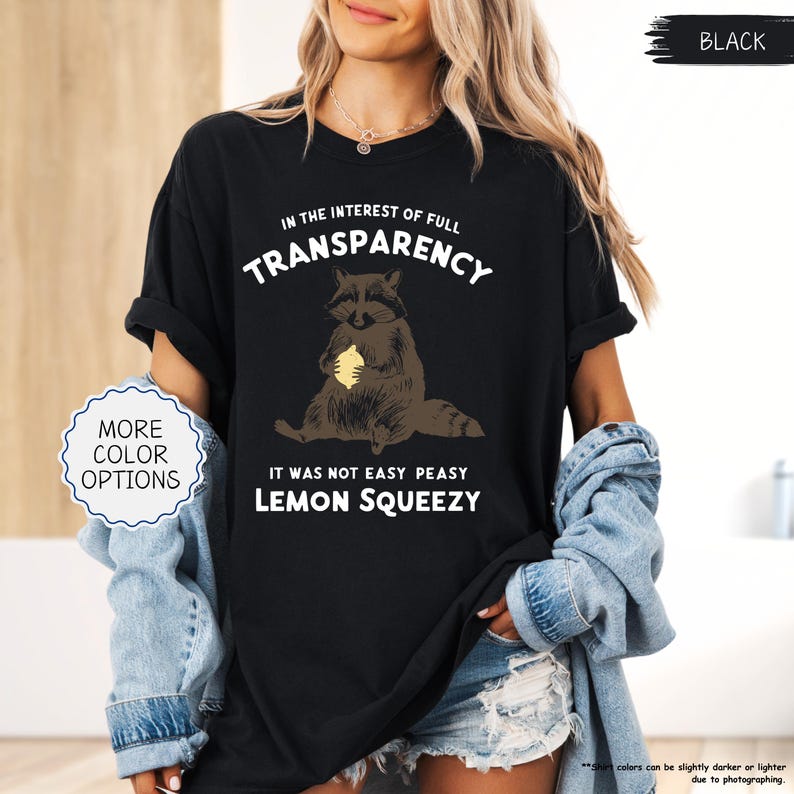 May include: Black t-shirt with a raccoon graphic holding a lemon. The text on the shirt reads "IN THE INTEREST OF FULL TRANSPARENCY", "IT WAS NOT EASY PEASY", and "LEMON SQUEEZY". The shirt also has a "MORE COLOR OPTIONS" button.