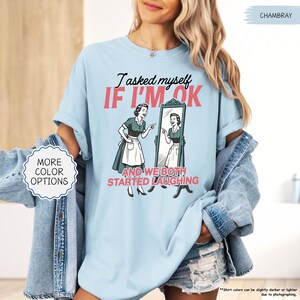 May include: Light blue t-shirt with the text "I asked myself IF I'M OK AND WE BOTH STARTED LAUGHING" and a retro illustration of a woman looking in a mirror. The shirt also has the words "MORE COLOR OPTIONS" on a white circle.