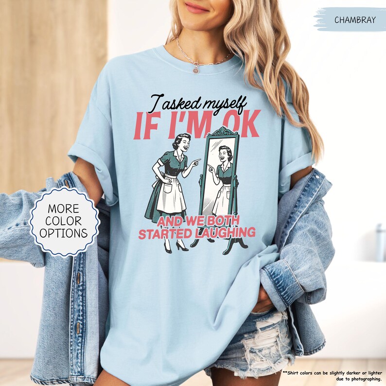 May include: Light blue t-shirt with the text "I asked myself IF I'M OK AND WE BOTH STARTED LAUGHING" and a retro illustration of a woman looking in a mirror. The shirt also has the words "MORE COLOR OPTIONS" on a white circle.