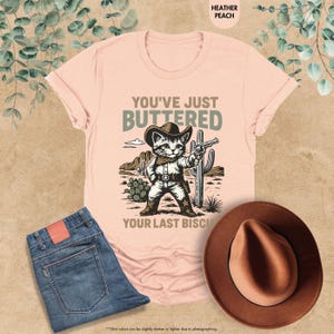 May include: Heather peach t-shirt featuring a cartoon cat in cowboy attire, holding a gun, with the words "YOU'VE JUST BUTTERED YOUR LAST BISCUIT". The image also shows blue jeans and a brown hat.