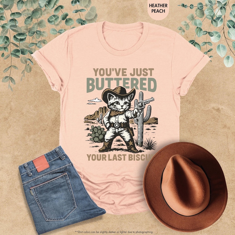 May include: Heather peach t-shirt featuring a cartoon cat in cowboy attire, holding a gun, with the words "YOU'VE JUST BUTTERED YOUR LAST BISCUIT". The image also shows blue jeans and a brown hat.