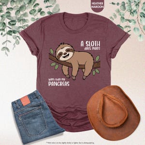K&ouml;nnte beinhalten: Weinrotes T-Shirt mit einem Faultier-Motiv. Das braune Faultier l&auml;chelt und ruht auf einem Ast mit gr&uuml;nen Bl&auml;ttern. Der Text lautet: "Ein Faultier arbeitet mehr als meine Bauchspeicheldr&uuml;se". Auch eine blaue Jeans und ein brauner Cowboyhut sind zu sehen.