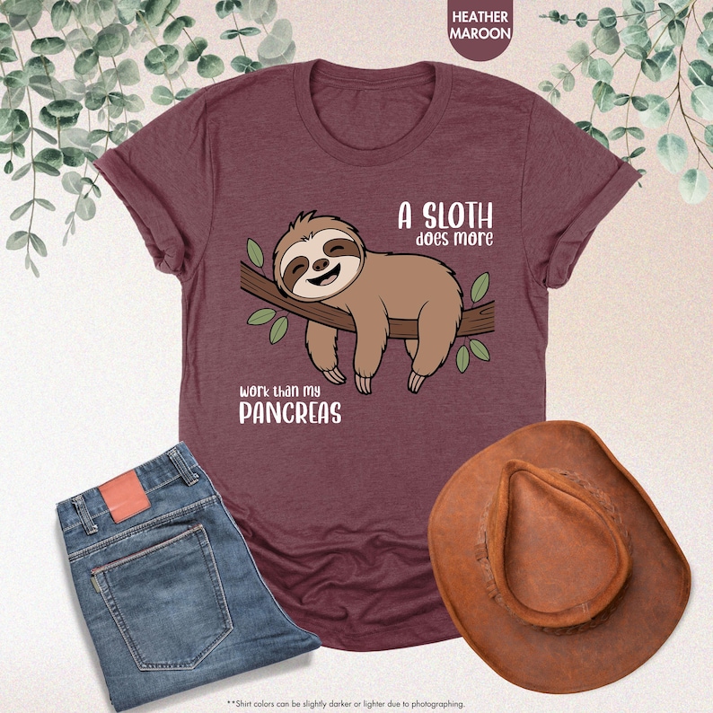 K&ouml;nnte beinhalten: Weinrotes T-Shirt mit einem Faultier-Motiv. Das braune Faultier l&auml;chelt und ruht auf einem Ast mit gr&uuml;nen Bl&auml;ttern. Der Text lautet: "Ein Faultier arbeitet mehr als meine Bauchspeicheldr&uuml;se". Auch eine blaue Jeans und ein brauner Cowboyhut sind zu sehen.