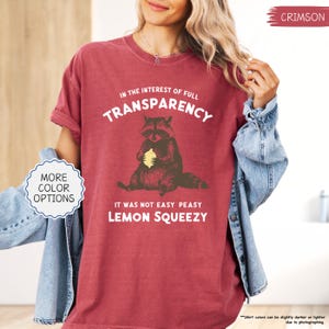 May include: Crimson-colored t-shirt with the text "IN THE INTEREST OF FULL TRANSPARENCY" and "IT WAS NOT EASY PEASY LEMON SQUEEZY". The shirt features a raccoon graphic holding a lemon. Additional color options are available.