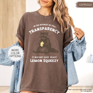 May include: Espresso-colored t-shirt with the text "IN THE INTEREST OF FULL TRANSPARENCY" and "IT WAS NOT EASY PEASY LEMON SQUEEZY". The shirt features a raccoon graphic holding a lemon. The shirt is a relaxed fit.