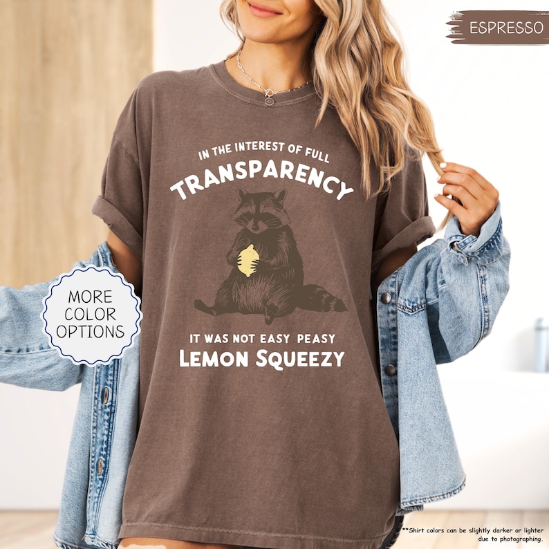 May include: Espresso-colored t-shirt with the text "IN THE INTEREST OF FULL TRANSPARENCY" and "IT WAS NOT EASY PEASY LEMON SQUEEZY". The shirt features a raccoon graphic holding a lemon. The shirt is a relaxed fit.