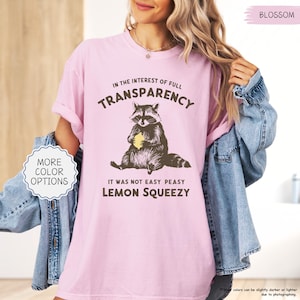 May include: A light pink t-shirt with the text "IN THE INTEREST OF FULL TRANSPARENCY" and "IT WAS NOT EASY PEASY LEMON SQUEEZY". The graphic features a raccoon holding a lemon. The shirt also has the word "BLOSSOM" printed on it.