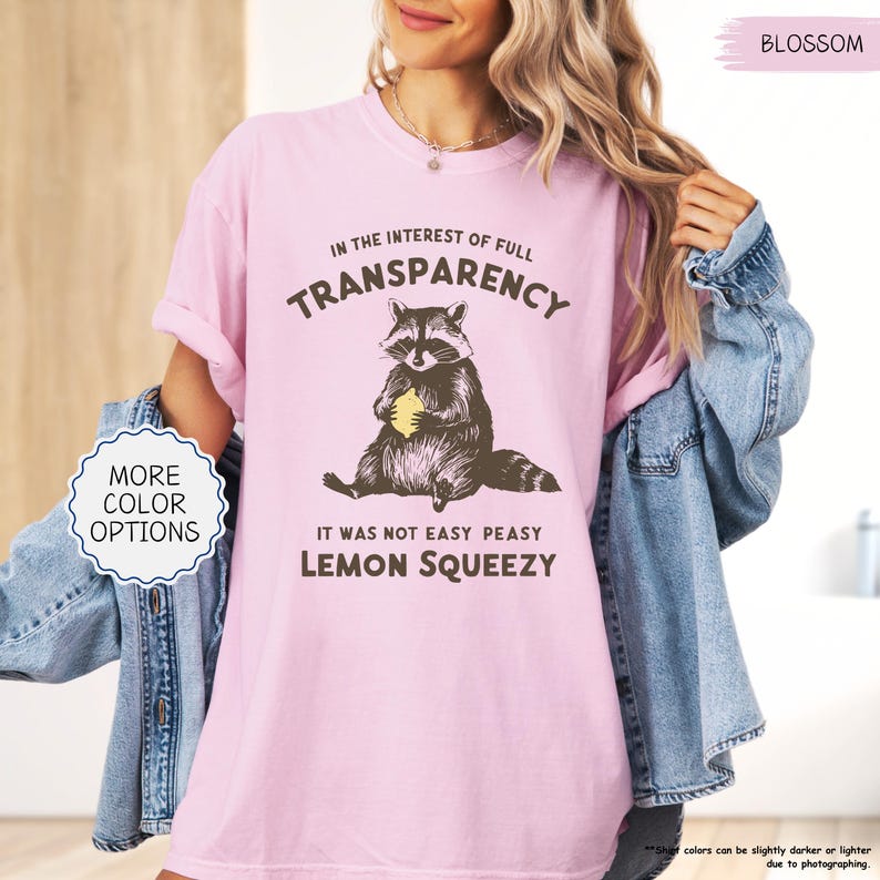 May include: A light pink t-shirt with the text "IN THE INTEREST OF FULL TRANSPARENCY" and "IT WAS NOT EASY PEASY LEMON SQUEEZY". The graphic features a raccoon holding a lemon. The shirt also has the word "BLOSSOM" printed on it.