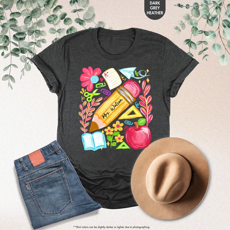 May include: Dark grey heather t-shirt with a colorful teacher-themed graphic. The design includes a large pencil with "Mrs. Watson" printed on it, surrounded by flowers, an apple, and school supplies. A pair of blue jeans and a tan hat are also in the image.