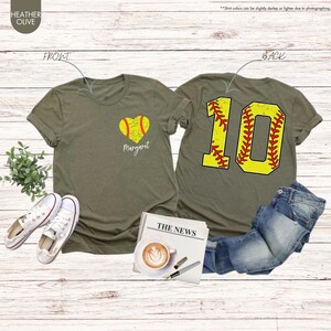 K&ouml;nnte beinhalten: Zwei olivgr&uuml;ne T-Shirts mit Softball-Motiv. Das vordere Shirt hat einen gelben Softball in Herzform mit dem Namen "Margaret" darunter. Das hintere Shirt hat eine gro&szlig;e gelbe Nummer "10" mit roten N&auml;hten, die einem Softball &auml;hnelt.