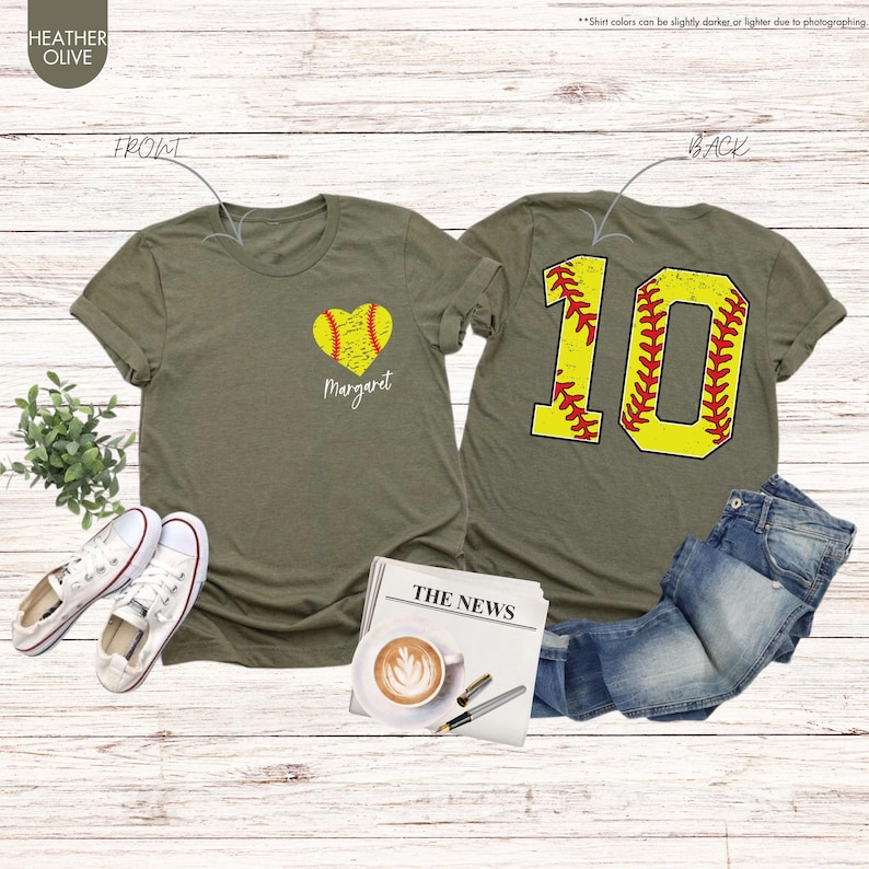 K&ouml;nnte beinhalten: Zwei olivgr&uuml;ne T-Shirts mit Softball-Motiv. Das vordere Shirt hat einen gelben Softball in Herzform mit dem Namen "Margaret" darunter. Das hintere Shirt hat eine gro&szlig;e gelbe Nummer "10" mit roten N&auml;hten, die einem Softball &auml;hnelt.