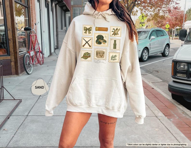 K&ouml;nnte beinhalten: Ein sandfarbener Kapuzenpullover mit einer Vordertasche und einer Kapuze mit Kordelzug. Der Kapuzenpullover zeigt eine Sammlung von Grafiken im Briefmarkenstil, darunter eine Schleife, ein Fernglas, eine Ente, gekreuzte Gewehre, ein Hund, eine Flasche, ein Hut, eine Kiste und ein Zweig Bl&auml;tter.