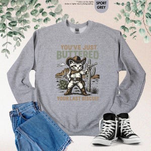 May include: Grey sweatshirt featuring a cowboy cat graphic with the text "YOU'VE JUST BUTTERED YOUR LAST BISCUIT". The design includes desert scenery and cactus. The sweatshirt is paired with blue jeans and black sneakers. The image is taken in natural light.