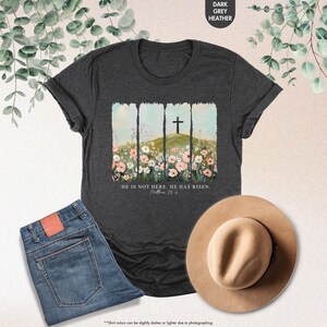 May include: A dark heather grey t-shirt with a floral design and a cross. The text "He is not here. He has risen. Matthew 28:6" is printed on the shirt.