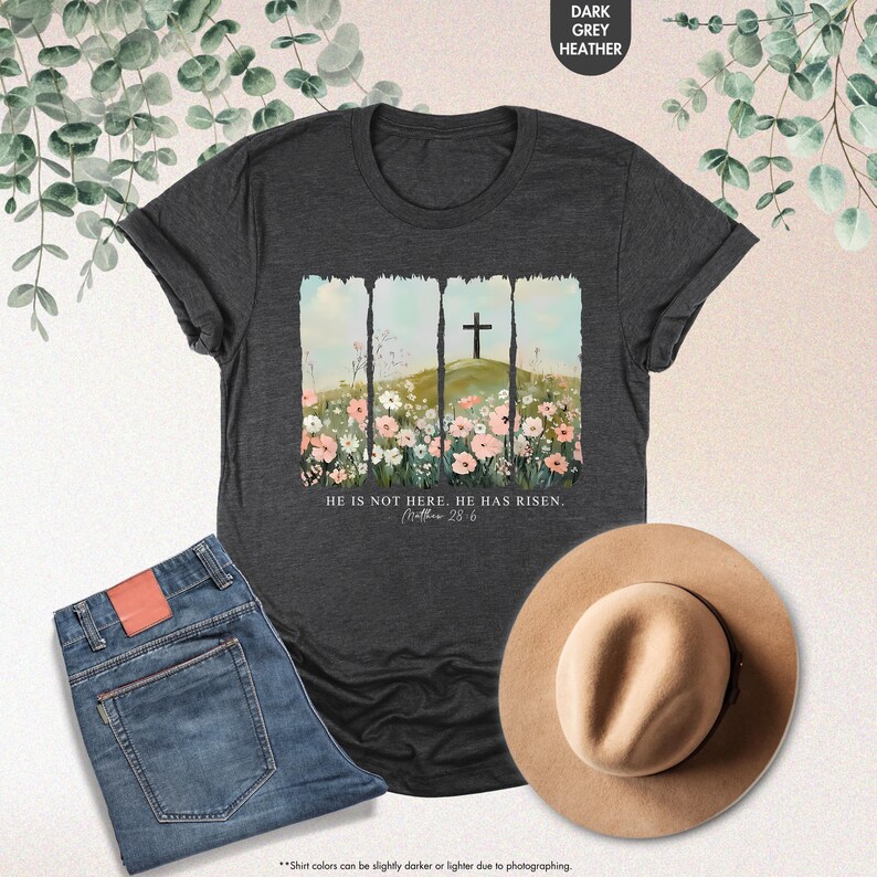 May include: A dark heather grey t-shirt with a floral design and a cross. The text "He is not here. He has risen. Matthew 28:6" is printed on the shirt.