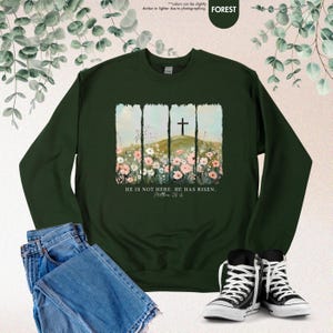 May include: A dark green sweatshirt with a floral design and a cross on a hill. The text "HE IS NOT HERE. HE HAS RISEN. (Matthew 28:6)" is printed on the sweatshirt.