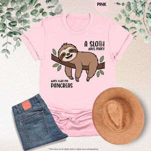 K&ouml;nnte beinhalten: Hellrosa T-Shirt mit einer Cartoon-Faultier-Illustration auf einem Ast und dem Text "A sloth does more work than my pancreas". Eine blaue Jeans und ein brauner Hut sind ebenfalls zu sehen.