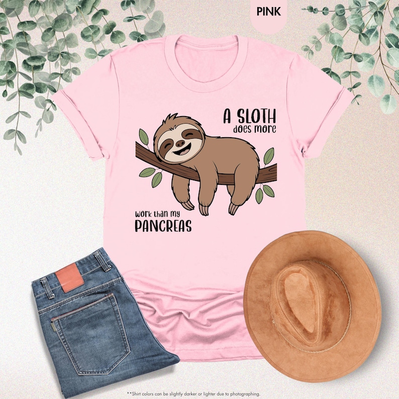 K&ouml;nnte beinhalten: Hellrosa T-Shirt mit einer Cartoon-Faultier-Illustration auf einem Ast und dem Text "A sloth does more work than my pancreas". Eine blaue Jeans und ein brauner Hut sind ebenfalls zu sehen.