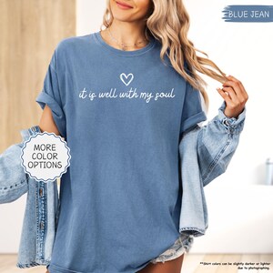 K&ouml;nnte beinhalten: Blaues Jeans-T-Shirt mit dem Schriftzug "it is well with my soul" in wei&szlig;er Schrift, &uuml;ber einem kleinen Herzen. Das Shirt hat hochgekrempelte &Auml;rmel und ist Teil einer Kollektion mit mehr Farboptionen.