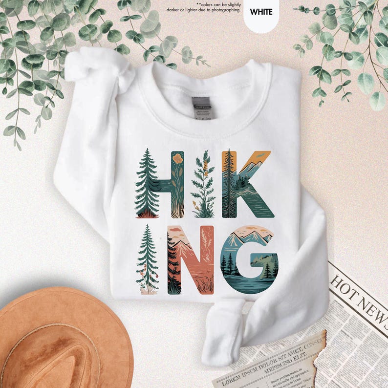 Pode incluir: Camisola branca com a palavra "HIKING" em letras grandes. Cada letra &eacute; preenchida com uma cena da natureza, incluindo &aacute;rvores, montanhas e um lago.
