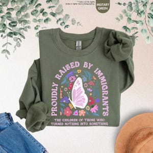 May include: Military green sweatshirt with the text "PROUDLY RAISED BY IMMIGRANTS" in a floral design, including a butterfly. The text "THE CHILDREN OF THOSE WHO TURNED NOTHING INTO SOMETHING" is also visible. The sweatshirt is displayed on a neutral background.