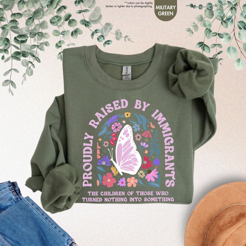 May include: Military green sweatshirt with the text "PROUDLY RAISED BY IMMIGRANTS" in a floral design, including a butterfly. The text "THE CHILDREN OF THOSE WHO TURNED NOTHING INTO SOMETHING" is also visible. The sweatshirt is displayed on a neutral background.