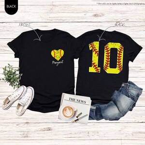 K&ouml;nnte beinhalten: Zwei schwarze T-Shirts mit Softball-Motiv. Das vordere Shirt hat ein Herz mit einem Softball darin und den Text "Margaret". Das hintere Shirt hat einen gelben Softball mit roten N&auml;hten und die Nummer "10" darauf.