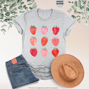May include: A gray t-shirt with a graphic of nine red and pink strawberries on a white background. The shirt is a relaxed fit and has a crew neck.
