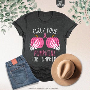 以下が含まれることがあります： 「CHECK YOUR PUMPKINS FOR LUMPKIN」の文字が入ったダークグレーのヘザーTシャツ。シャツには、骸骨の手とピンクのリボンが付いた2つのピンクのかぼちゃのグラフィックが描かれています。青いジーンズとベージュの帽子も表示されています。