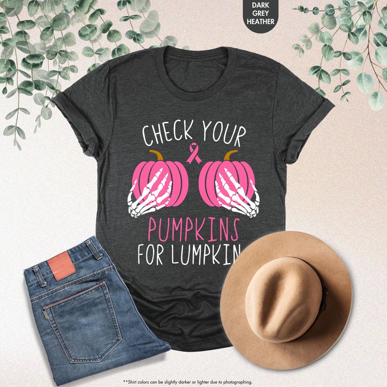 以下が含まれることがあります： 「CHECK YOUR PUMPKINS FOR LUMPKIN」の文字が入ったダークグレーのヘザーTシャツ。シャツには、骸骨の手とピンクのリボンが付いた2つのピンクのかぼちゃのグラフィックが描かれています。青いジーンズとベージュの帽子も表示されています。