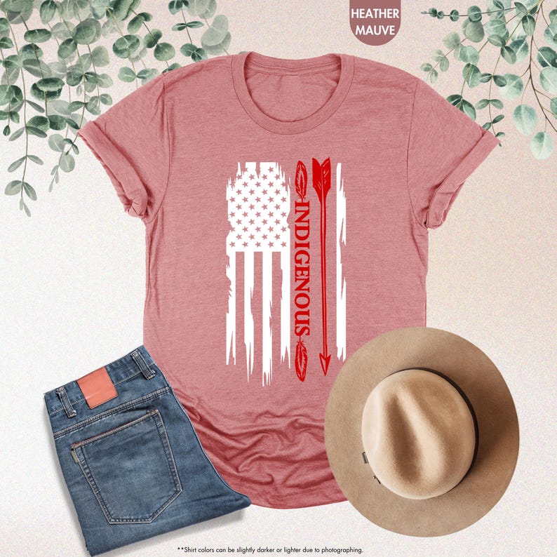 May include: A heather mauve t-shirt with a white distressed American flag design and the word "INDIGENOUS" in red lettering. The flag is flanked by two red arrows.
