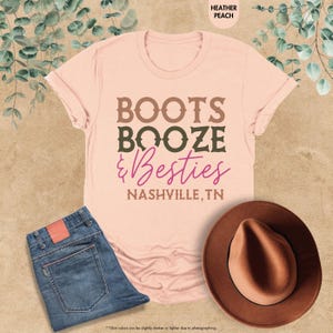Pu&ograve; includere: Una maglietta color pesca con le parole "BOOTS, BOOZE, & Besties, NASHVILLE, TN" in lettere marroni e viola. La maglietta &egrave; abbinata a jeans blu e un cappello marrone.