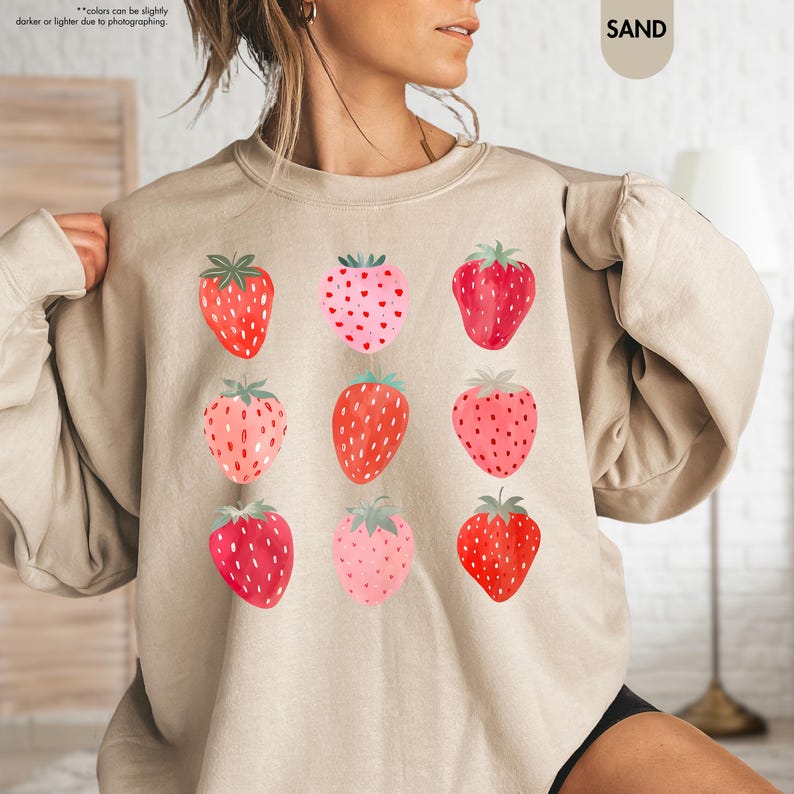 May include: A beige sweatshirt with a graphic print of nine pink and red strawberries. The text "SAND" is printed in the top right corner of the image.